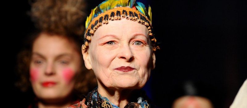 An older woman, fashion designer Vivienne Westwood, with a feathered headdress in front of a group of people.