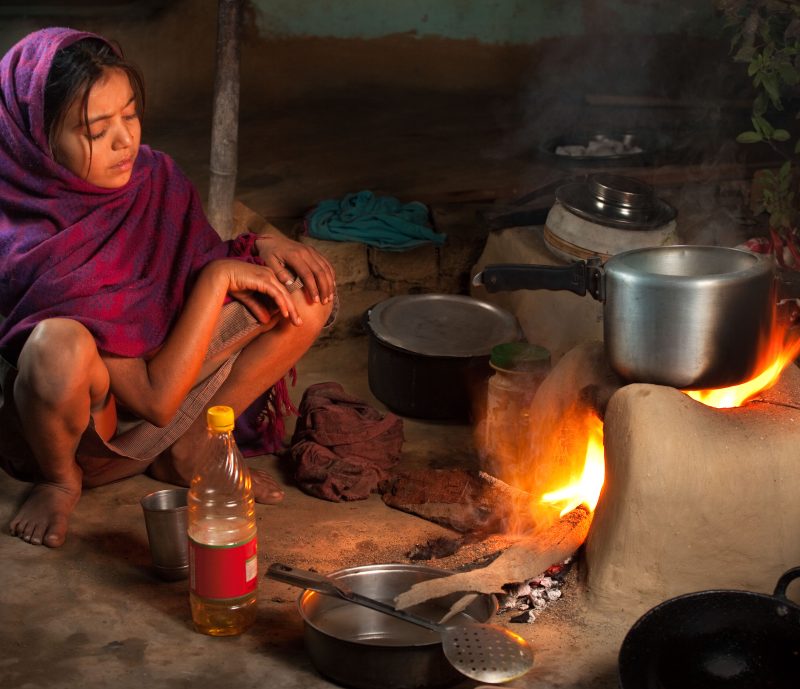 A person cooking on a traditional clay stove, covered with a purple shawl, surrounded by cooking utensils and a plastic bottle, embodies the contrast in the age of Net-Zero Obsession.