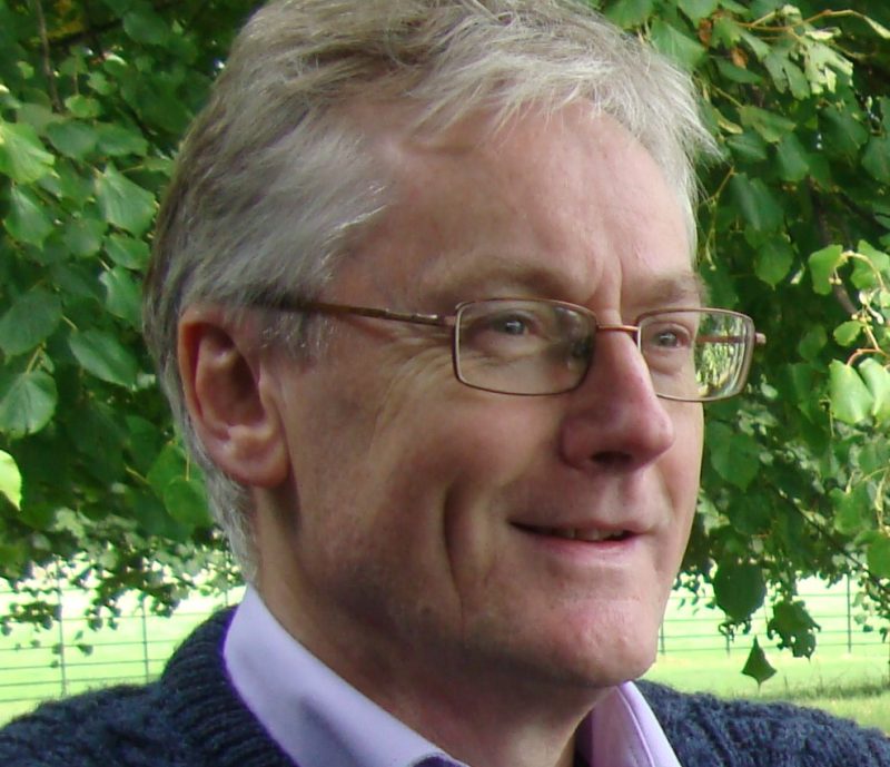 Amidst green leaves, Mike Hulme stands outdoors with his signature glasses and grey hair, wearing a blue sweater over a purple shirt. He is poised for an interview, adding depth to the Profiles in Sustainability series.