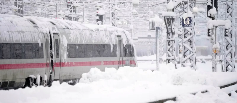 A train covered in snow moves along tracks at a station during heavy snowfall, with snow piling on the ground and overhead wires.