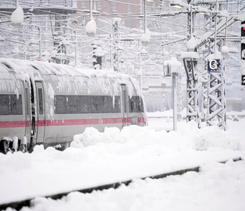 A train covered in snow moves along tracks at a station during heavy snowfall, with snow piling on the ground and overhead wires.