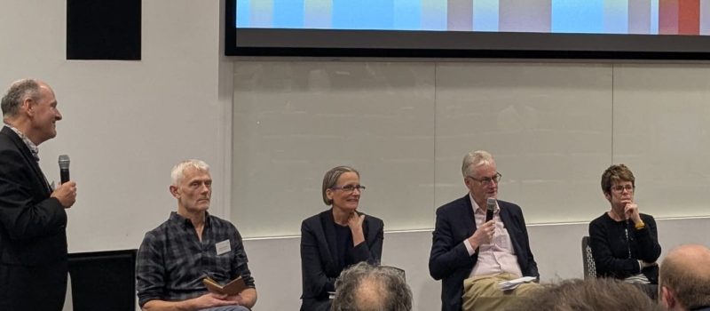 Five people participate in a panel discussion on "What I Would Have Done Differently"; one stands holding a microphone while the other four are seated with microphones and notes in front of a presentation screen.