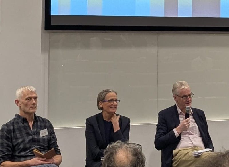 Five people participate in a panel discussion on "What I Would Have Done Differently"; one stands holding a microphone while the other four are seated with microphones and notes in front of a presentation screen.