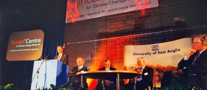 A panel of five people sit on stage under banners for the Tyndall Centre and University of East Anglia, as one man speaks at a podium about climate challenges like floods at COP.