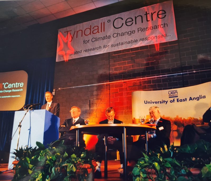 A panel of five people sit on stage under banners for the Tyndall Centre and University of East Anglia, as one man speaks at a podium about climate challenges like floods at COP.