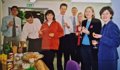 A group of eight adults in business attire stand around a table with snacks and drinks, smiling at the camera in an office setting, perhaps after discussing climate topics like COP failure or recent Tyndall Centre research on floods.