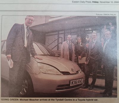A group of men in suits stand around a silver Toyota hybrid car outside a building, one man resting his hand on the car's bonnet, discussing recent floods and the ongoing research by the Tyndall Centre.