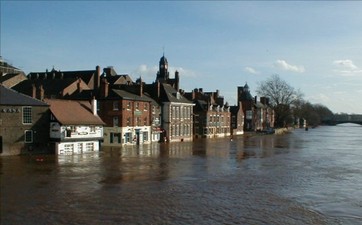 Row of brick buildings partially flooded by a river, with water covering the lower floors—a stark reminder of the increasing floods highlighted by the Tyndall Centre—set against a clear blue sky.